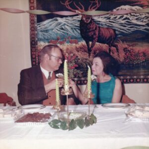 Brewer wedding reception James Moran Troutman wearing a dark suit and Maria Troutman wearing a teal dress during their (first) wedding reception. They smile at each other as they hold drinks while seated in front of a white tablecloth-covered table with lit candles in holders and a wedding cake. A decorative tapestry hangs behind them.
