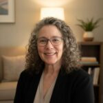 Debra Feinberg, LCSW with dark, curly shoulder-length hair smiling while wearing glasses and a dark blazer over a light-colored blouse.