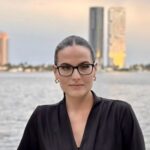 Dr. Carolina Estevez in front of a riverside city skyline wearing black glasses and a black shirt, her hair tied back.