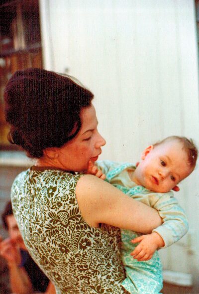 Maria&toddlerMichelleAug1974-front Maria Troutman with dark hair wearing a patterned sleeveless top holding Michelle Troutman (as a toddler) wearing light blue clothing. Michelle is looking to her left while her mother, in profile, looks at her.